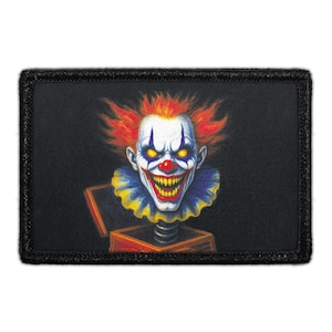May include: A rectangular patch featuring a graphic illustration of a sinister clown with bright orange hair, glowing yellow eyes, and a wide grin with sharp teeth. The clown wears a blue and yellow ruffled collar and emerges from a red jack-in-the-box.
