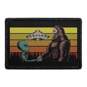 May include: A rectangular embroidered patch featuring a graphic of a green alien and a brown Bigfoot shaking hands. Above them is a silver flying saucer. The background displays a sunset with horizontal stripes in yellow, orange, and red.