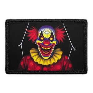 May include: A rectangular patch featuring a menacing clown with red hair, a yellow ruffled collar, and a red polka-dotted shirt. The clown has a wide, toothy grin, yellow eyes, and is set against a black background.