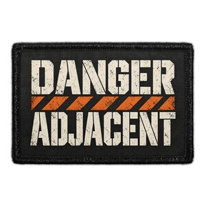 May include: A rectangular black patch with a distressed white text graphic that reads DANGER ADJACENT. The design features a horizontal orange stripe with black diagonal hazard lines separating the two words, all set against a dark background.
