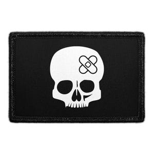 May include: A rectangular black patch featuring a white graphic of a human skull. The skull has a white adhesive bandage with a central square detail positioned on its forehead. The patch has a stitched border and a flat, fabric-like texture.