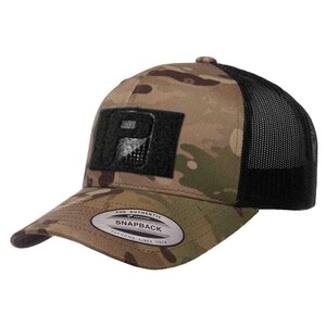 May include: A camouflage print snapback cap with a black mesh back and a black rectangular patch on the front featuring a stylized letter P. The brim displays a silver sticker with the text The Authentic Yupoong Snapback Yupoong Since 1974.