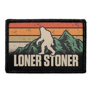 May include: A rectangular embroidered patch with a black border featuring a graphic of a beige Bigfoot silhouette walking in front of green mountains and a sunset sky with horizontal stripes of orange, yellow, and beige. The text LONER STONER is printed in beige capital letters at the bottom.
