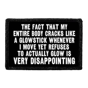 May include: A rectangular black patch with a stitched border featuring white text that reads: THE FACT THAT MY ENTIRE BODY CRACKS LIKE A GLOWSTICK WHENEVER I MOVE YET REFUSES TO ACTUALLY GLOW IS VERY DISAPPOINTING. The patch has a clean, bold font.