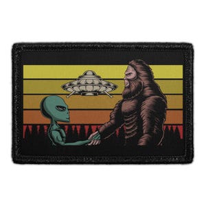 May include: A rectangular embroidered patch featuring a graphic of a green alien and a brown Bigfoot shaking hands. Above them is a silver flying saucer. The background displays a sunset with horizontal stripes in yellow, orange, and red.