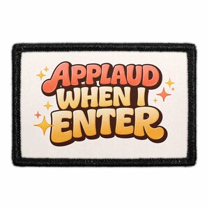 May include: A rectangular patch with the text "APPLAUD WHEN I ENTER" in orange and yellow, with star accents. The patch has a black border and a cream-colored background.