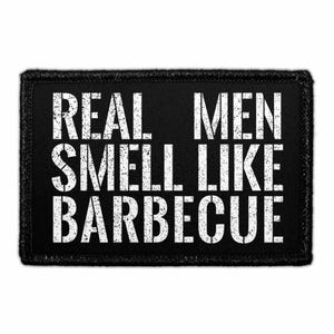 May include: A rectangular black patch with a white distressed-style font that reads REAL MEN SMELL LIKE BARBECUE. The text is arranged in three lines with a bold, sans-serif typeface, and the patch features a stitched border around the edge.