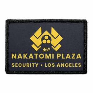 May include: A rectangular patch with a black border and a navy blue background. The patch features gold text and a gold graphic. The text reads "NAKATOMI PLAZA SECURITY • LOS ANGELES". There is also Japanese text and a graphic.