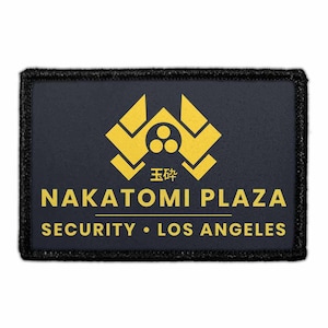 May include: A rectangular black patch with a gold embroidered logo featuring a stylized emblem above the text NAKATOMI PLAZA, the Japanese characters 玉砕, and the words SECURITY • LOS ANGELES printed in a bold, sans-serif gold font.