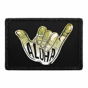 May include: A rectangular black patch featuring a white shaka hand gesture graphic filled with a green and cream tropical leaf pattern. The word ALOHA is printed in bold black capital letters across the palm of the hand graphic on the patch.