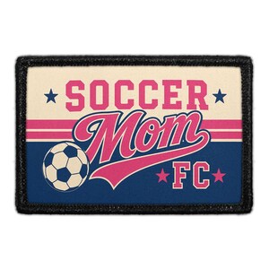May include: A rectangular patch with a black border featuring a cream top section and a dark blue bottom section. The text SOCCER MOM FC is printed in pink with a soccer ball graphic on the left. Two pink stripes and four blue stars are included.