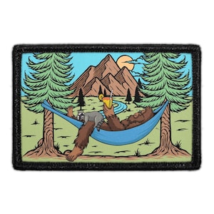 May include: A rectangular embroidered patch featuring a brown, furry creature resting in a blue hammock between two pine trees. The creature holds a yellow drink with a straw, while a small raccoon sits on its lap. Mountains and a sun appear in the background.