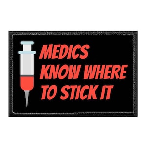 May include: A rectangular black patch with a stitched border featuring a graphic of a syringe with a red liquid inside. The text MEDICS KNOW WHERE TO STICK IT is printed in bold red capital letters to the right of the syringe illustration.