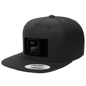 May include: A solid black snapback cap featuring a structured crown and a flat brim. A rectangular black patch is centered on the front panel, displaying a dark gray graphic logo of a stylized letter P with a dotted texture pattern on the lower half.