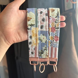 Puede incluir: Tres llaveros de tela floral para muñeca con herrajes de oro rosa. Los llaveros presentan una variedad de estampados florales en tonos blancos, amarillos, rosas y azules. Cada llavero tiene forma rectangular con cierre y anilla metálicos.