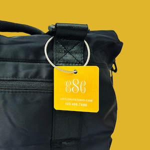 May include: A black tote bag with a yellow tag attached to it. The tag has the number 886 and the text "LITTLEGOOTSSTUDIO.COM 123 456 7890".