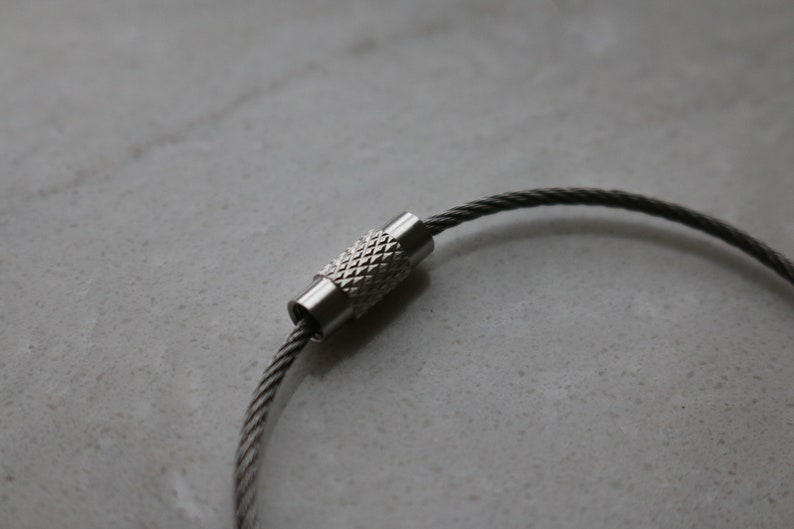 May include: A silver key chain with a braided cable and a silver clasp. The clasp has a textured design.