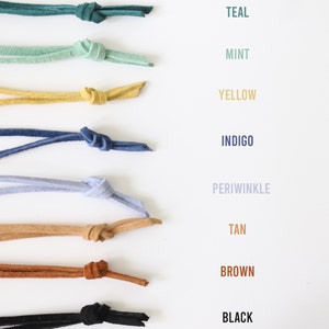May include: A collection of eight leather cords in various colors, including teal, mint, yellow, indigo, periwinkle, tan, brown, and black. Each cord is tied in a knot and arranged vertically on a white background.