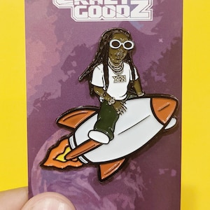 Puede incluir: Un pin de esmalte blanco y naranja con una ilustración de dibujos animados de una persona con rastas que lleva gafas de sol y una camisa blanca con el texto "YFN" en ella, montando un cohete sobre un fondo morado y blanco. El pin tiene el texto "CRAZY GOODZ" en la parte superior y "CRAZYGOODZ.COM" en la parte inferior.