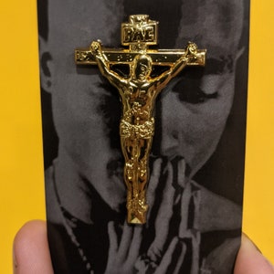 May include: Gold-colored metal pin of a person on a cross with the word "RAVE" on the top of the cross.