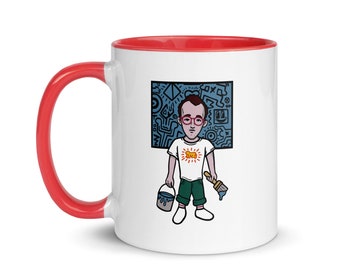Peut inclure: Une tasse en céramique blanche avec un bord et une poignée rouges. La tasse présente une illustration de dessin animé d'une personne portant des lunettes tenant un pinceau et un pot de peinture devant un fond abstrait bleu et noir. La personne porte une chemise blanche avec un graphique de soleil jaune et un pantalon vert.