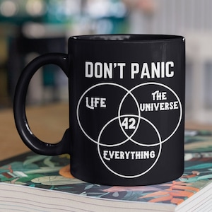 May include: Black ceramic mug with a white design that says "Don't Panic" and features a Venn diagram with the words "Life", "The Universe", and "Everything" and the number 42 in the centre.