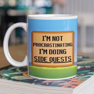 May include: White ceramic mug with a blue and green pixelated design. The mug features the text "I'm not procrastinating I'm doing side quests".