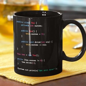 May include: Black coffee mug featuring a humorous programming code design. The code is written in a light blue, pink, and white font. The code reads: "public class Tea { private int ounces; public Tea() { this.ounces = 11; } public void drink(int sip) { this.ounces -= sip; } } Tea tea = new Tea(); while (tea.ounces != 0) { tea.drink(1); } System.out.println("Get shit done!");"