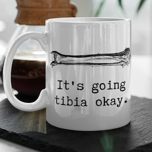 May include: White ceramic mug with a black and white illustration of a tibia bone. The mug has the text "It's going tibia okay" in black. The mug is on a black surface.
