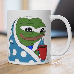 May include: White ceramic mug with a green frog wearing a blue and white polka dot blanket and holding a red mug of coffee.