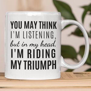 May include: White ceramic mug with black text that reads "You may think I'm listening, but in my head, I'm riding my triumph."