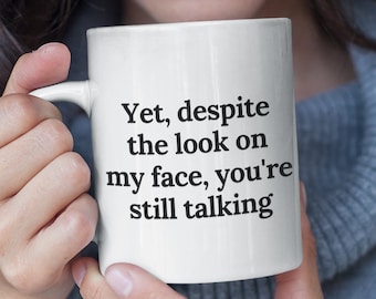 May include: White ceramic mug with black text that reads "Yet, despite the look on my face, you're still talking".