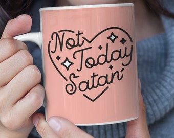 May include: A peach-colored ceramic mug with the phrase "Not Today Satan" in black script and a heart design. The mug is held by a person wearing a gray sweater and glasses.