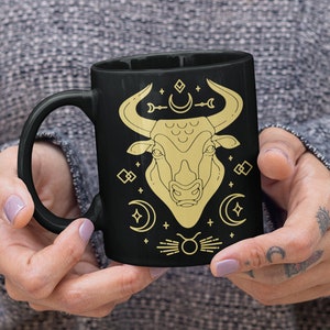 May include: Black ceramic mug with a gold outline of a bull's head and astrological symbols. The bull's head is surrounded by crescent moons, stars, and a symbol of the zodiac sign Taurus.