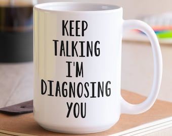 May include: White ceramic coffee mug with black text that reads "Keep Talking I'm Diagnosing You". The mug is sitting on a brown notebook.