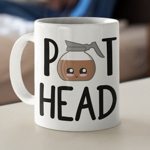 May include: White ceramic coffee mug with the words "POT HEAD" in black block letters. The letter "O" in "POT" is replaced with a cartoon coffee pot with a smiling face. The mug has a large, curved handle.