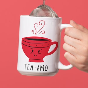 May include: White ceramic mug with a red cartoon teacup with a smiling face and the text "TEA-AMO". A silver tea infuser is hanging from the mug.