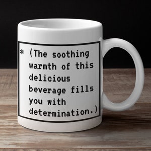 May include: White ceramic mug with a black border around a text graphic that reads "* (The soothing warmth of this delicious beverage fills you with determination.)"