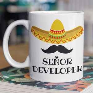 Puede incluir: Taza de café de cerámica blanca con un bigote negro y un sombrero amarillo con detalles rojos y verdes. El texto "Señor Developer" está impreso en negro debajo del sombrero.