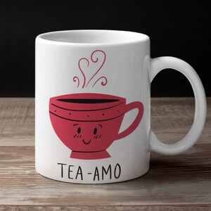 May include: White ceramic mug with a red teacup with a smiling face and steam coming out of it. The text "TEA-AMO" is printed below the teacup.