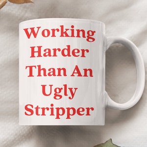 May include: White ceramic coffee mug with red text that reads "Working Harder Than An Ugly Stripper".