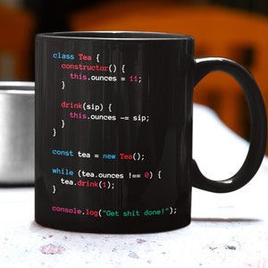 Könnte beinhalten: Schwarze Keramik-Tasse mit Programmiercode-Design. Der Code enthält "class Tea", "constructor" und "console.log("Get shit done!")". Die Tasse steht auf einer weißen Oberfläche, mit einer silbernen Tasse im Hintergrund.