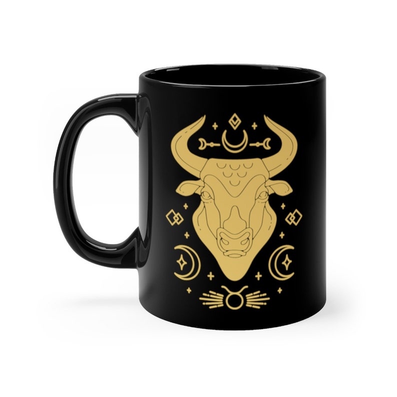 May include: Black ceramic mug with a gold outline of a bull's head and astrological symbols. The bull's head is surrounded by crescent moons, stars, and a Taurus zodiac symbol.