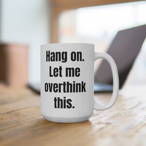 K&ouml;nnte beinhalten: Eine wei&szlig;e Keramik-Kaffeetasse mit schwarzem Text, der "Hang on. Let me overthink this." lautet.