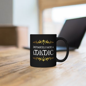May include: Black coffee mug with gold accents and the text "Definitely not a Mimic" in white.