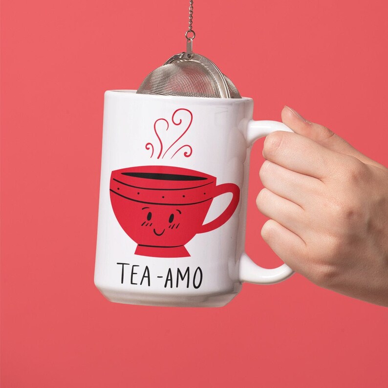 May include: White ceramic mug with a red cartoon teacup with a smiling face and the text "TEA-AMO". A silver tea infuser is hanging from the mug.