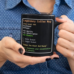 May include: Black coffee mug with a green and white design that looks like a video game interface. The mug says "Legendary Coffee Mug" and has stats like "+69 Stamina" and "Keeps You Alert And Ready".