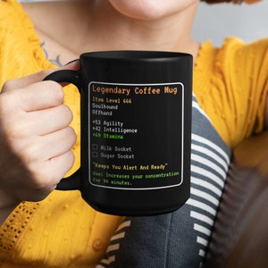 May include: Black coffee mug with a green and orange design that looks like a video game character stats screen. The text reads "Legendary Coffee Mug", "Item Level 666", "Soulbound", "Offhand", "+53 Agility", "+42 Intelligence", "+69 Stamina", "Milk Socket", "Sugar Socket", "Keeps You Alert And Ready", "Use: Increases your concentration for 90 minutes."