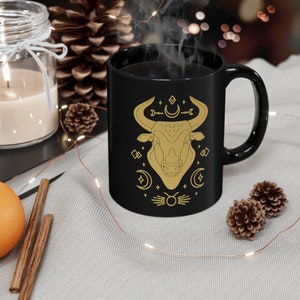 May include: Black ceramic mug with a gold design of a bull's head with crescent moons and stars. The mug has a white interior and a handle.
