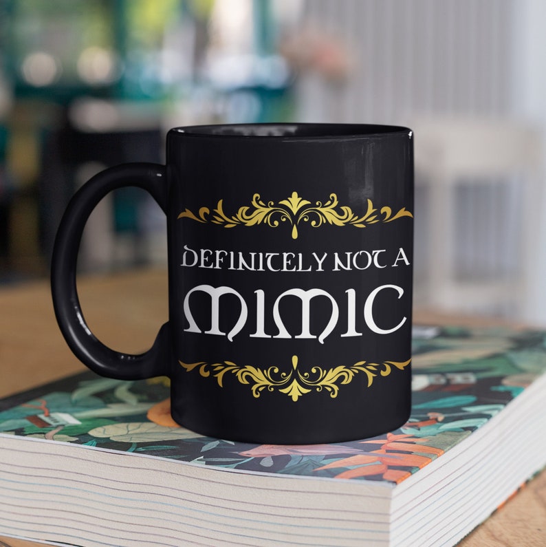 May include: Black ceramic coffee mug with a gold design and the text "Definitely not a mimic" in white lettering.